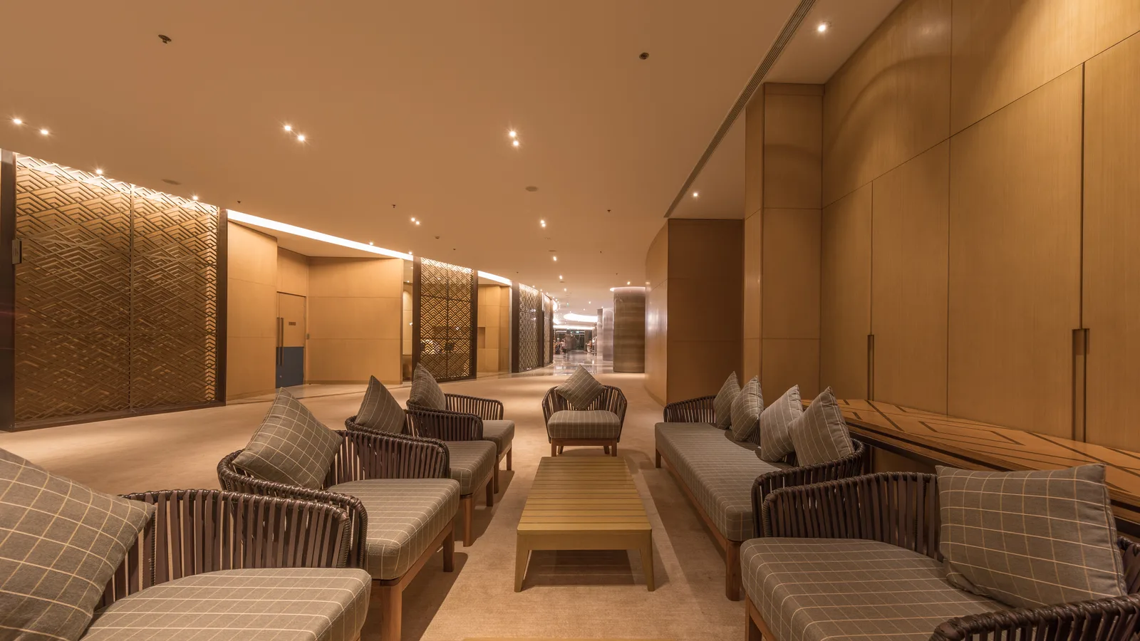A hotel club lounge corridor with paired rattan armchairs, low tables and warm wood panelling running the length of the room
