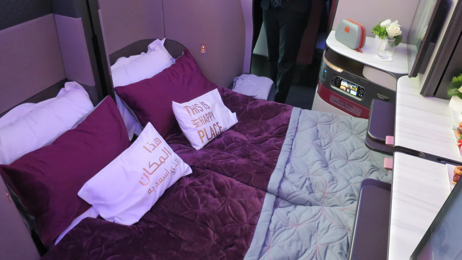 A Qatar Airways Qsuite business class double bed made up with burgundy bedding, twin pillows and a folded blanket beside the side console