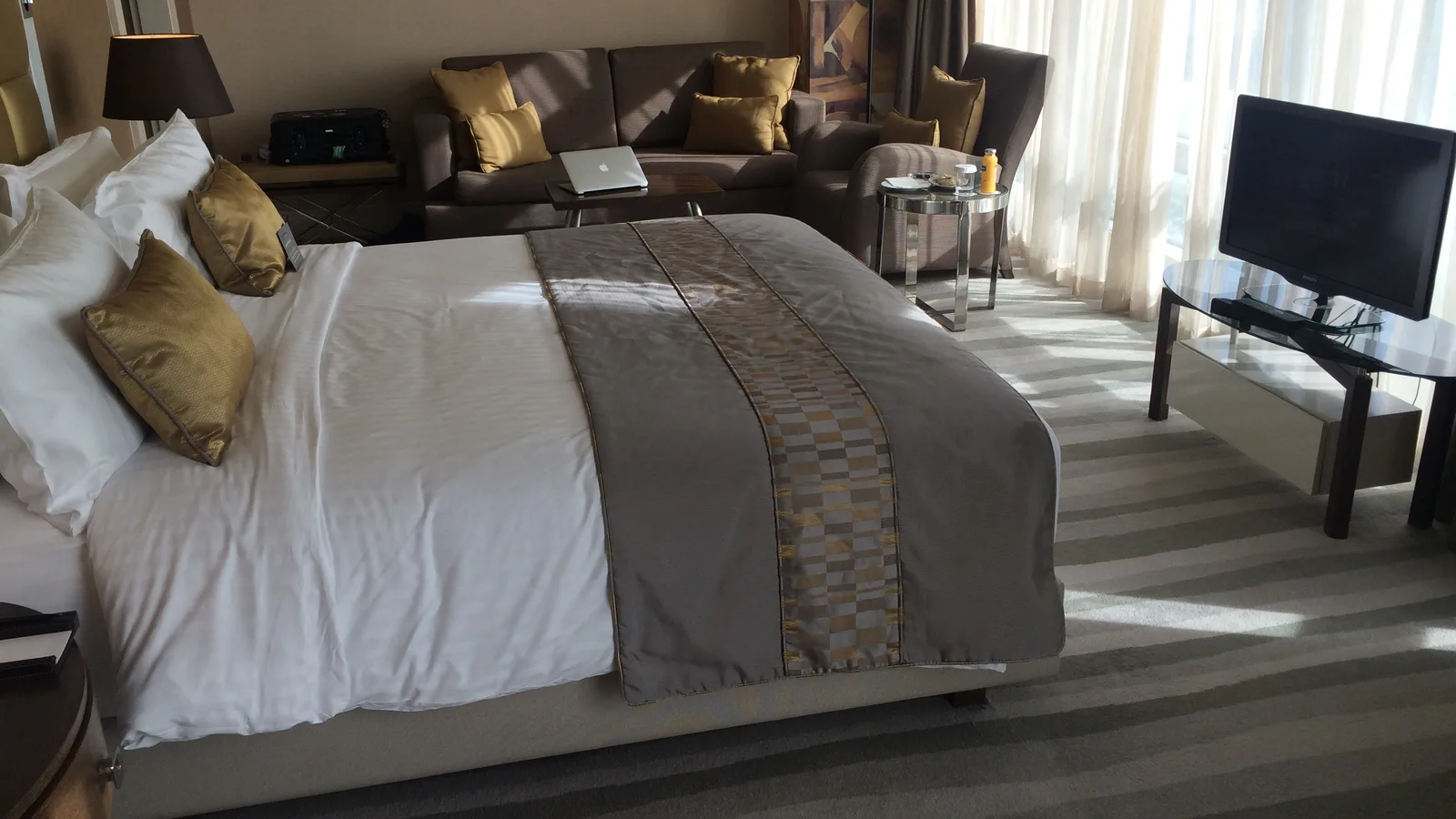 An upscale hotel guest room with a king bed, gold accent cushions, a lounge sofa and floor-to-ceiling window drapes