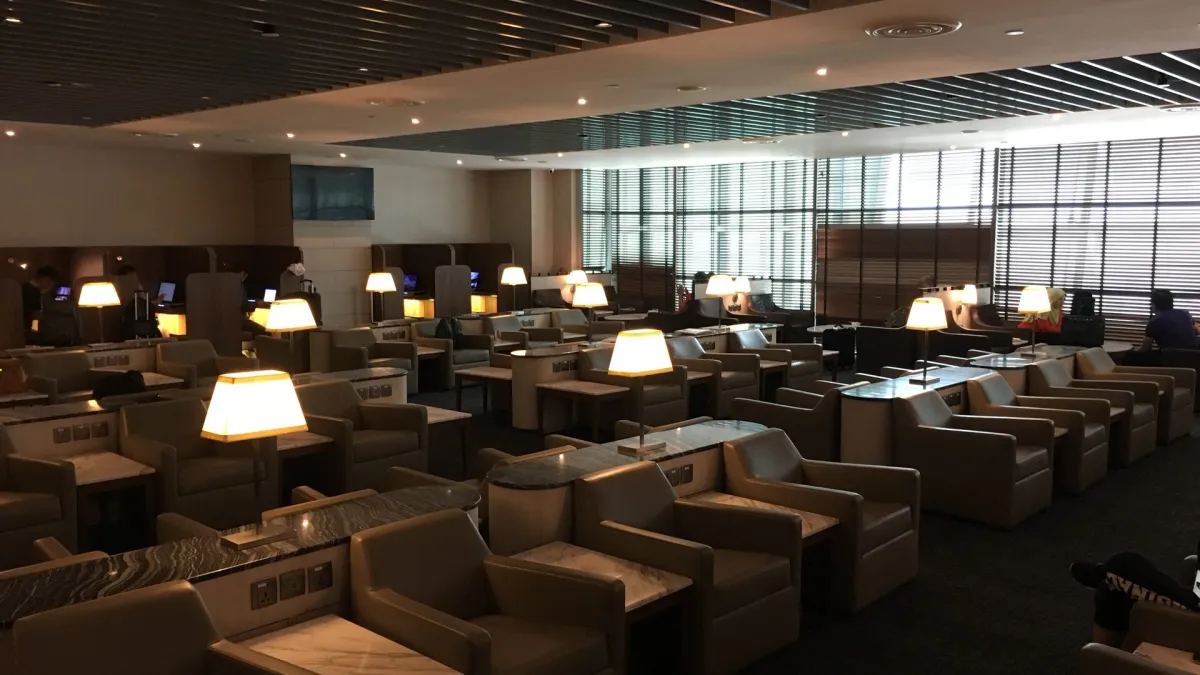 A wide, quiet airport business lounge with rows of tan leather armchairs, individual table lamps and slatted blinds along the window wall